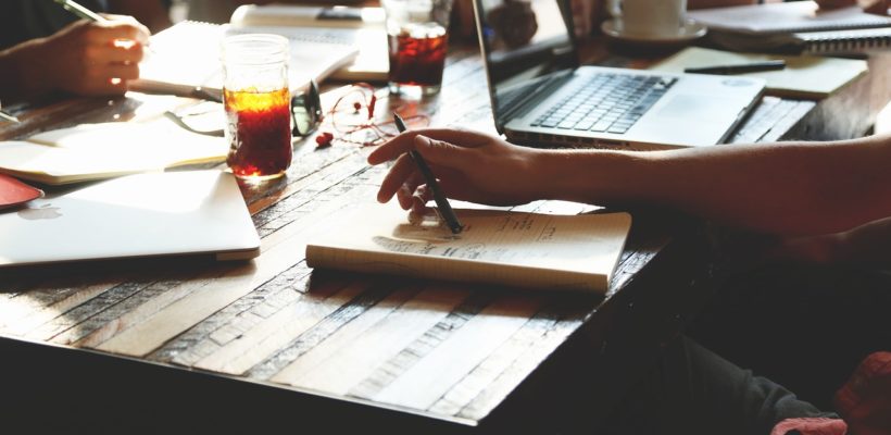 A hand over a notebook with a computer in the background